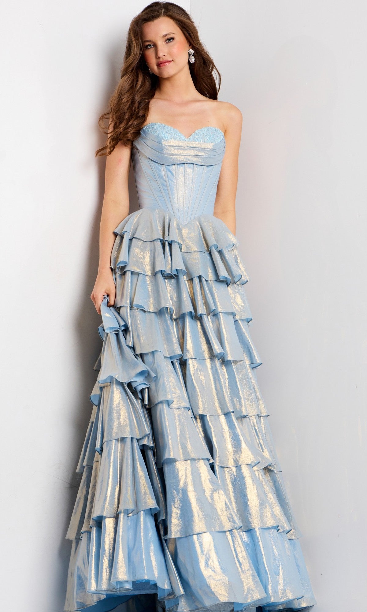 A woman stands against a plain light background wearing the Jovani 47030 long prom dress—a strapless, light blue gown with a sweetheart neckline, corset bodice, and multiple ruffled, tiered layers.